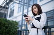© WMSTUDIO - Young woman in stylish attire using smartphone while standing outdoors near modern building with lush greenery on a bright sunny day