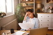 © Stockphotodirectors - A woman with a concerned expression examines multiple papers related to insurance problems in her well-lit home office. She sits at a wooden desk cluttered with documents and a laptop.
