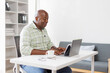 © David - Senior businessman sitting alone in his home office and working on laptop computer. Smiling black man using laptop in living room. Businessman using phone laptop at home.