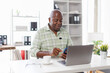 © David - Senior businessman sitting alone in his home office and working on laptop computer. Smiling black man using laptop in living room. Businessman using phone laptop at home.