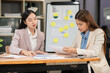 © Thitisak - Two Asian businesswomen holding graph papers are sitting and talking happily while working.