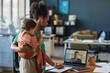 © DragonImages - Person holding baby while working on laptop and participating in video call, emphasizing work-from-home lifestyle and balancing responsibilities