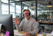 © Centric  - Cheerful businessman wearing headphones, waving and smiling at computer during a video call in a bright, modern office setting
