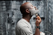 © TrueFrame Collective - Black man with beard shaving in front of mirror in modern bathroom, using shaving cream and razor. Man is taking care of grooming and hygiene without looking at camera