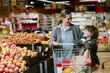 © AnnaStills - Smiling woman pushing shopping cart with happy child embracing her in grocery store produce section filled with fruits and vegetables
