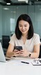 © Anwar Fauzan - Middle-aged Asian business woman manager ceo using cell phone mobile app, laptop. Smiling mature businesswoman entrepreneur holding smartphone sitting in office working online on gadget, copy space
