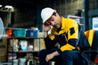 © Ekkasit A Siam - Caucasian adult male warehouse worker in uniform and helmet resting on chair wiping sweat with tired face after working long shift in industrial storage zone surrounded by work-related items