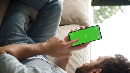 Relaxed phone overlay, man lying on couch using smartphone with green screen showing blank mobile interface for chroma video app mockup or digital product demo