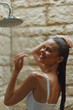 © SHOTPRIME STUDIO - smiling woman enjoying outdoor shower with wet hair, natural stone background, warm lighting, relaxed mood, casual white swimsuit, summer freshness