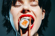 © VICHIZH - A young woman with short black hair passionately eating sushi using chopsticks, showcasing excitement and joy in her expression against a dark backdrop.
