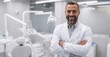 © Stock Pix - Smiling dentist in a white coat, arms crossed, in a modern dental office. He is confident and welcoming to his patients.