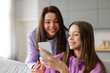 © Home-stock - Happy teenage girl showing cellphone to her mother, sharing interesting content, funny photos or videos, sitting on sofa at home