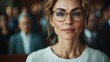 ©  zikakant - A confident woman with curly hair and glasses gazes seriously at the camera, surrounded by a blurred audience in a formal setting, emphasizing focus and poise.