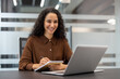 © Liubomir - A woman with curly hair smiles while writing notes during an online meeting in the office. Her laptop sits open in front of her.