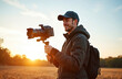 © miss irine - Young man films with camera, gimbal at sunrise. Videographer content creator makes video. Smiling guy with cap, backpack shoots media. Light lens, film equipment, outdoors.