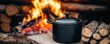 © Thanagorn - A black cast iron pot sits on a wooden surface near a campfire with stacked logs in the background.