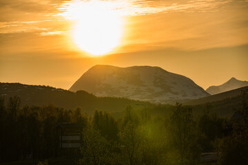 Wall Mural - Bright golden sun setting behind snowy mountain peaks in Sweden, casting warm hues over trees and the landscape, creating a stunning evening panorama filled with natural beauty.