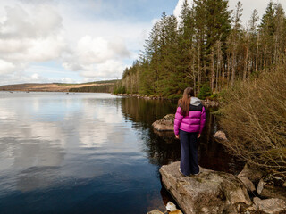 Wall Mural - Girl tourist enjoys view on a lake with cloudy sky reflection. Travel and tourism concept. Water is calm and green bushes and trees growing on the lake bank. Calm relaxed mood. Voyage and adventure.