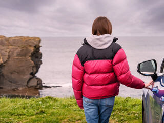 Wall Mural - A woman in a red jacket stands next to a blue car in a parking lot and looking at a stunning nature scene with cliff, rough stone coastline and cloudy sky. Travel and tourism. West of Ireland.
