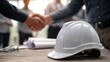 © CStock - A construction helmet rests on a table during a handshake agreement, symbolizing partnership and collaboration.