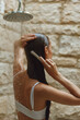 © SHOTPRIME STUDIO - Woman combing wet hair under shower water with natural stone wall background, calm and refreshing morning routine emphasizing self-care and hygiene.