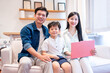© Q88 - A smiling family of three is sitting on a couch together The woman is holding a pink laptop