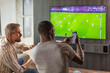 © ty - Two friends holding smartphone with sports bets app while watching football match at home in living room