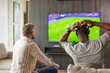 © ty - Two friends holding smartphone with sports bets app while watching football match at home in living room