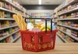 © Thecreator T - Red shopping basket filled with groceries on a wooden table in a supermarket aisle
