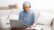 © Prostock-studio - Happy black woman in hijab wearing headphones, watching webinar on laptop, taking notes at home. Positive female student in traditional headscarf participating in online educational conference
