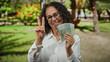 © Krakenimages.com - Woman smiling in outdoor park holding chilean pesos with glasses and gesturing peace sign.