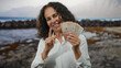 © Krakenimages.com - Woman holding colombian pesos smiles by the seaside, showcasing banknotes with the beach and waves in the background.