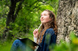 © Sonya Monshtein - Portrait of young woman with curly hair holding pen to chin looking up and dreaming writing in notebook relaxing resting alone dressed in blue shirt and jeans sitting near rock in forest outdoors