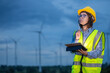© BESTIMAGE - A woman technician wearing a yellow vest and a hard hat points to a tablet. She is standing in front of a field of wind turbines