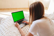 © fotofabrika - Woman sits on a cozy bed working on a laptop with a green screen during daylight in a bright room