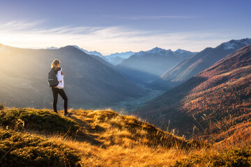 Wall Mural - Young woman with backpack standing on grassy mountain ridge with view of valley with high mountain peaks during golden hour sunset in autumn. Hiking in Italy, Livigno. Girl in Alps in fall. Trekking