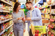 © Prostock-studio - Healthy Eating. Young Smiling Couple Buying Groceries In Supermarket Store Indoors. Two Buyers Holding Glass Jar Of Sauce And Checking Label, Choosing Food Products, Standing With Shopping Trolley