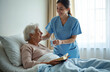 © Vadym - Care worker offers water elderly woman in geriatric hospice. Nurse and patient smiling. Senior lady reads book in bed. Healthcare support. Geriatric care assistance concept.