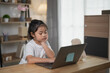 © WMSTUDIO - Young child engaged in online learning with laptop at home, focusing on study materials while sitting at a wooden desk with a toy bear beside her