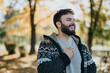 © qunica.com - A cheerful man in a warm, patterned sweater stands amidst autumn trees, laughing freely. The sunlit background and fallen leaves capture the essence of an enjoyable, relaxed outdoor moment.