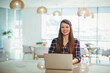 © WavebreakMediaMicro - Woman wearing plaid shirt working on laptop at modern office with wooden desk, pendant lamps, plant