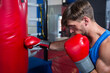 © wavebreak3 - Muscular boxer punching heavy bag near ring ropes on padded gym floor wearing red gloves