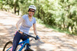 © wavebreak3 - Female cyclist riding blue bicycle along sunlit dirt trail in woods wearing helmet and sunglasses