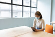 © WavebreakMediaMicro - Asian woman wearing face mask and pale top sitting at desk holding tablet near grid window