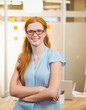 © WavebreakMediaMicro - Red-haired woman standing crossing arms in office by glass partition with sticky notes and laptop