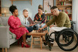 © Seventyfour - Diverse group of young adults and middle aged man with disability in wheelchair playing chess together in living room, Black woman and Caucasian man watching, multiethnic friends celebrating birthday