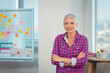 © WavebreakMediaMicro - Senior woman standing with arms crossed in office near whiteboard covered in sticky notes and desk