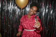 © Seventyfour - Portrait of young adult Black woman smiling and holding champagne glass while celebrating birthday in front of metallic fringe backdrop, gold balloon visible in hand