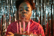 © Seventyfour - Portrait of Black woman blowing out candles on birthday cake, celebrating birthday in front of metallic backdrop, holding cake with lit candles, wearing earrings