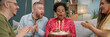 © Seventyfour - Website header shot of Black woman blowing out candles on birthday cake while celebrating holiday with diverse group of young adults clapping, while sitting on sofa in living room